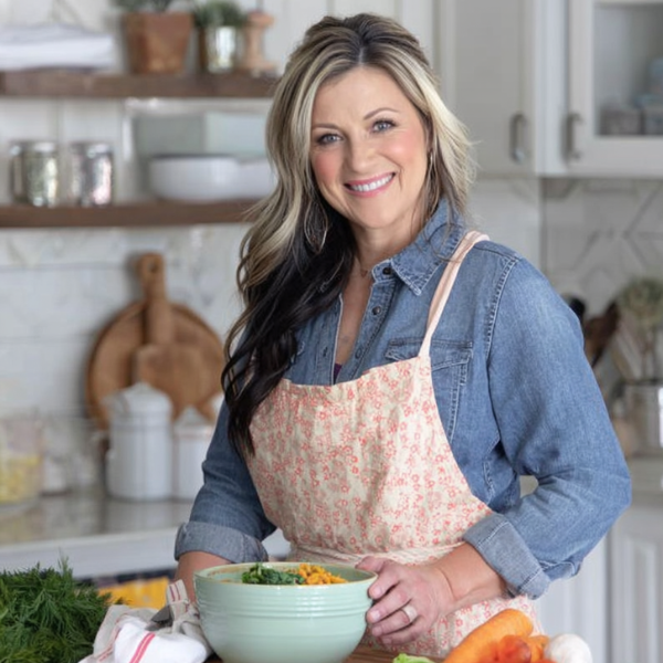 Paula Dena smiling in her kitchen, wearing a floral apron and preparing a fresh homemade dish